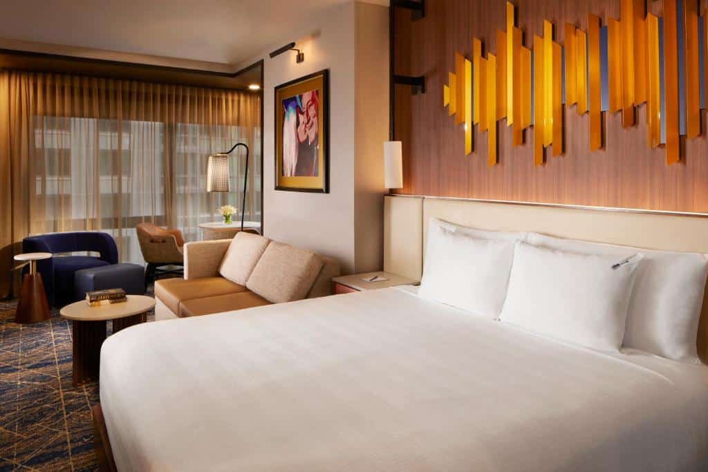 Quarto do Hard Rock hotel com uma cama de casal grande, um sofá, uma pequena mesa de centro e uma decoração elegante. As janelas grandes estão cobertas por cortinas escuras, e há um quadro colorido na parede da Lady Gaga beijando o rosto de Tony Bennet.