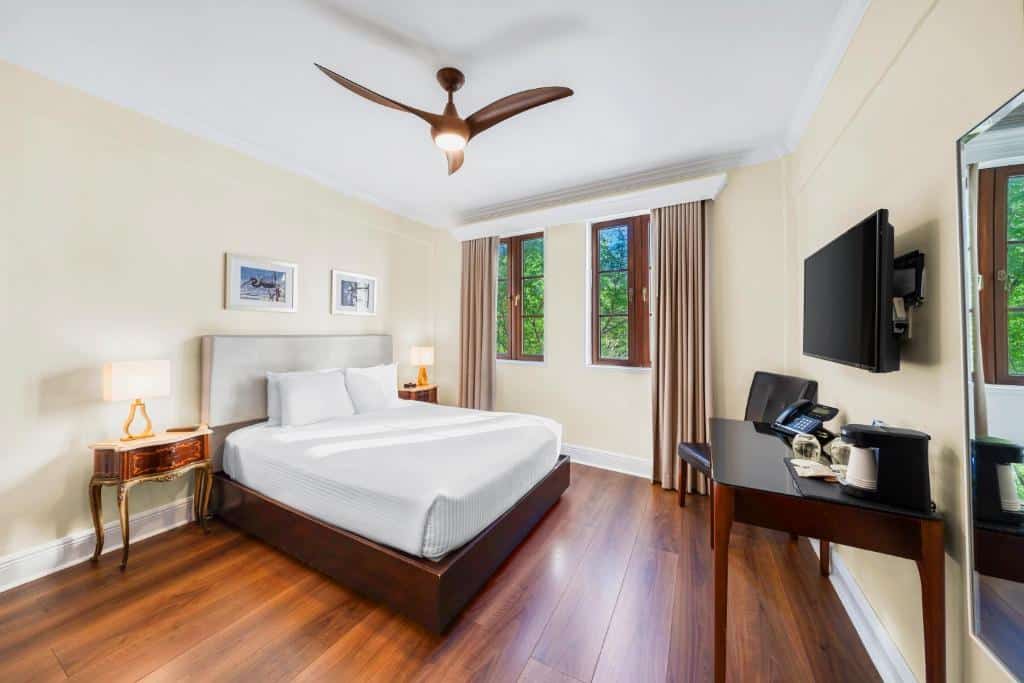 Quarto do Hotel St. Michel com cama de casal do lado esquerdo com uma comoda em cada lado da cama com luminária em frente a cama uma mesa com máquina de café e telefone e preso na parede uma TV do lado esquerdo da mesa uma cadeira e no teto no centro um ventilador de teto. Representa melhores hotéis em Miami.