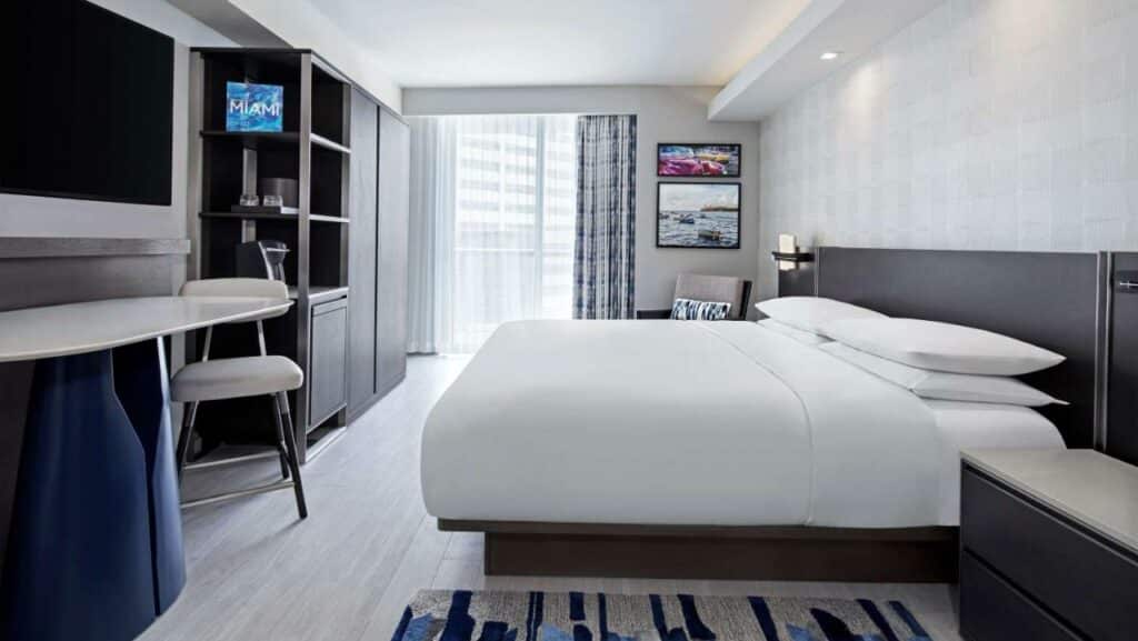 Quarto do Hyatt Centric Brickell Miami com uma cama grande, escrivaninha e cadeira. Arte nas paredes, uma pequena prateleira e uma janela com cortinas. Representa hotéis em Brickell.
