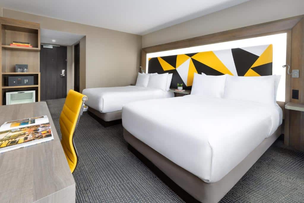 Quarto do M Social hotel com duas camas de casal, uma mesa de trabalho com cadeira amarela, e uma decoração moderna com cabeceira geométrica em amarelo, preto e branco. Este é um dos hotéis em Nova York.