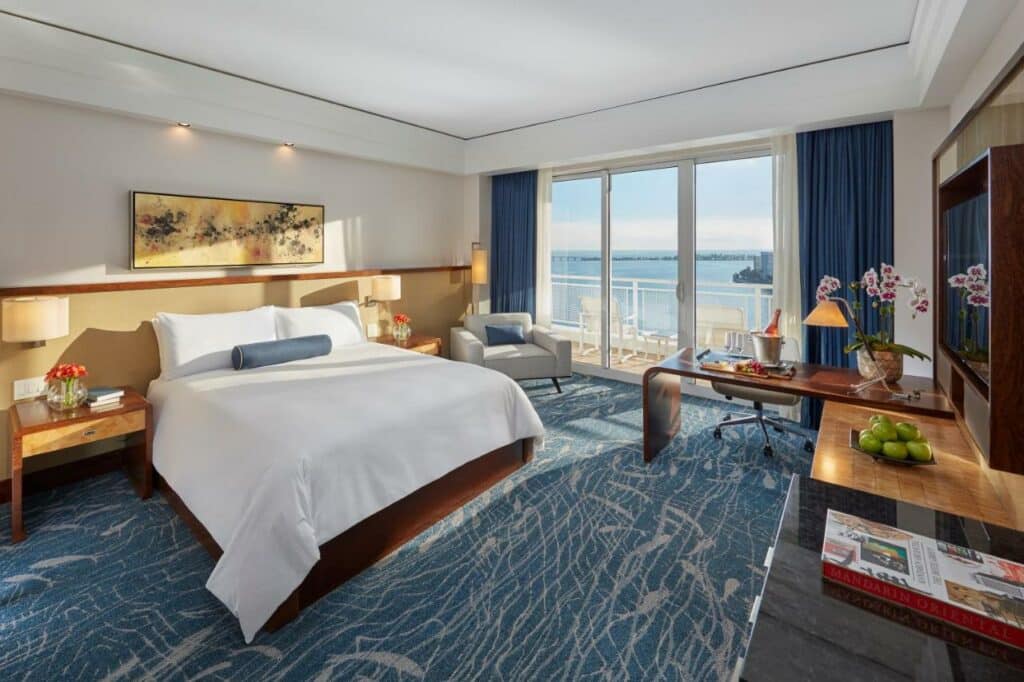 Quarto do Mandarin Oriental com uma cama grande, escrivaninha, poltrona e uma janela grande com vista para um corpo d'água. Decoração azul e branca por toda parte. Representa hotéis bem localizados em Miami.