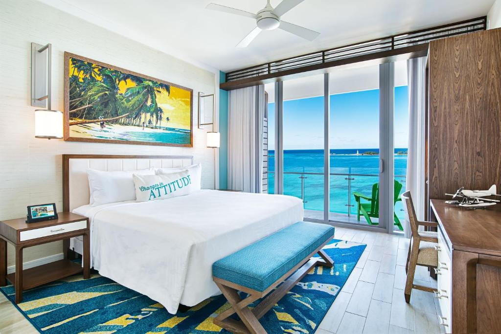 Quarto do Margaritaville Beach Resort Nassau com uma cama de casal com travesseiros e cobertas. Nos pés da cama tem um banco, do lado direito da cama tem uma mesa de cabeceira e em baixo um tapete. Do lado esquerdo tem uma porta grande de vidro que dá acesso a varanda do quarto com uma cadeira e vista para o mar.
