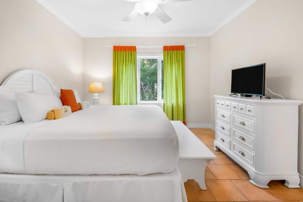 Quarto do Pineapple Fields que mostra uma cama alta de casal com um banco no pé da cama. Em frente ao banco tem uma cômoda com uma tv em cima. Ao fundo do quarto tem uma janela de vidro com cortinas.