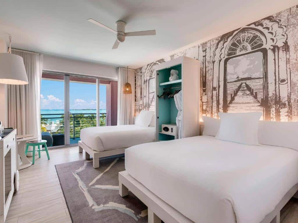 Quarto do SLS at Baha Mar que mostra duas camas, uma cama de casal e uma cama de solteiro. As duas estão separadas por um guarda-roupa. No quarto tem um tapete, uma cômoda e uma mesinha com um banco redondo. Do lado da cama de solteiro tem uma porta grande de vidro com cortinas que dá acesso a varanda do quarto com vista para a paisagem.