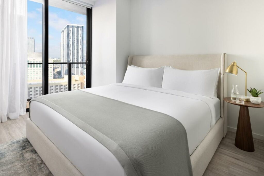 Quarto do The Elser Hotel Miami – An All-Suite Hotel com cama de casal do lado direito da imagem com uma mesa com luminária do lado esquerdo da cama. Representa melhores hotéis em Miami.