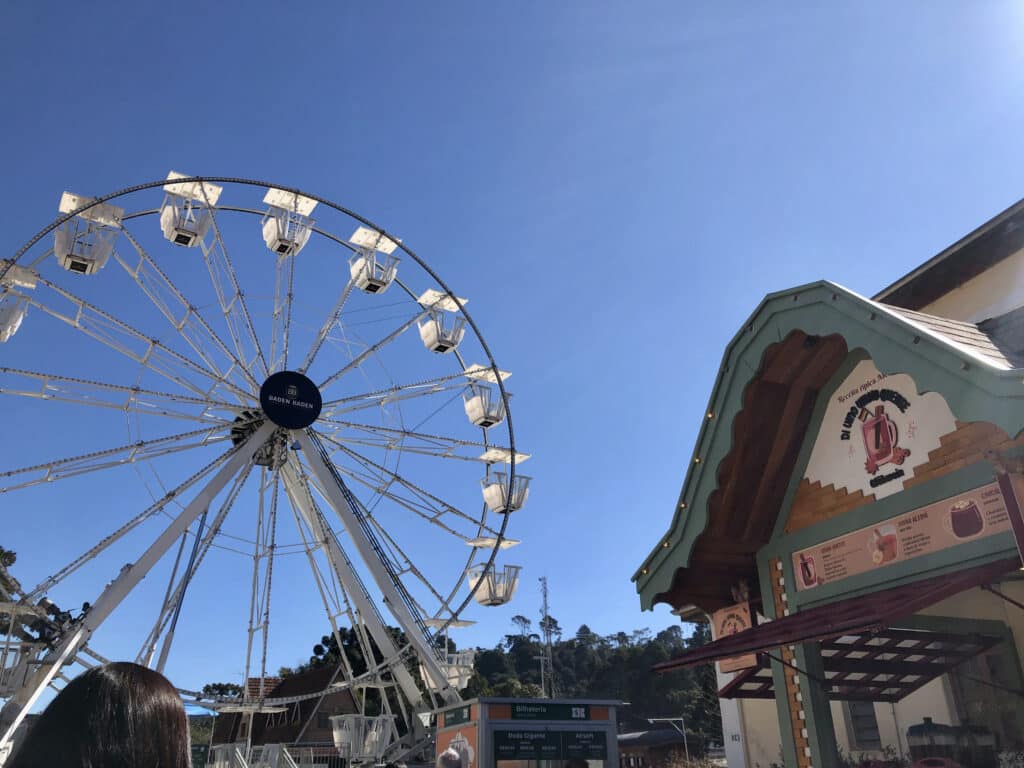 Foto da roda gigante no Parque Capivari. Ela está na esquerda, e na direita há uma loja de bebidas como chocolate quente e chá. O céu está azul.