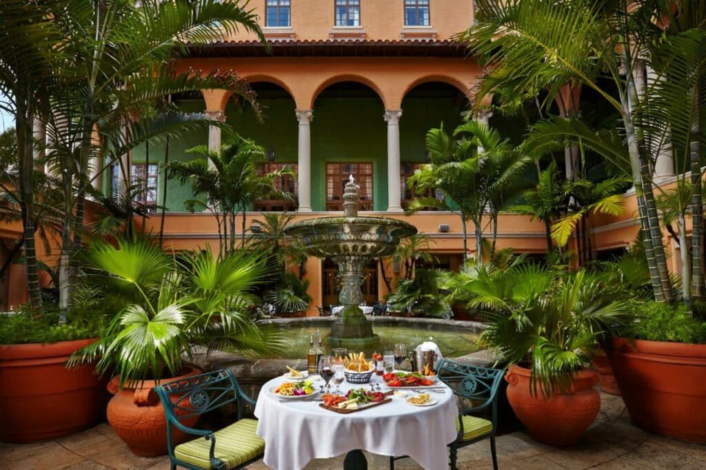 Ambiente para refeições ao ar livre do Biltmore Hotel com toalha de mesa branca, comida e bebidas em um pátio com fonte e plantas tropicais.