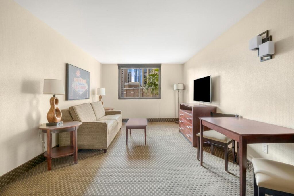 Sala de estar do The Carriage House. Um sofá está do lado esquerdo, ao lado de duas mesinhas com abajures. No meio está uma mesa de centro. Em frente ao sofá e a mesa está a TV. No canto direito da imagem há uma mesa retangular e duas cadeiras.