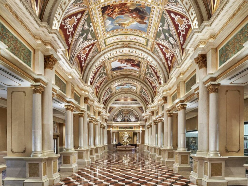 Lobby no The Venetian. O espaço é bem grande e remete a arquitetura italiana. O teto tem formato mais arredondado e é todo coberto com pinturas clássicas. Pilastras estão dos dois lados e formam um caminho central. O chão também é todo decorado com formatos diferentes.