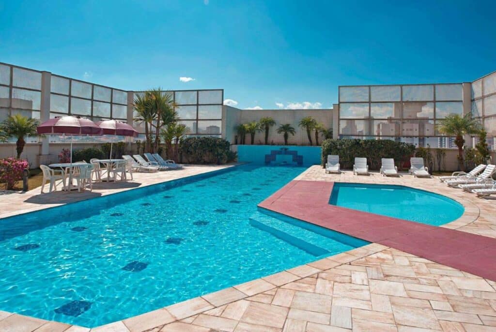 Uma piscina grande e uma piscina pequena ao ar livre no Hotel Numaz Brás. Ao redor cadeiras de tomar sol e guarda-sóis.