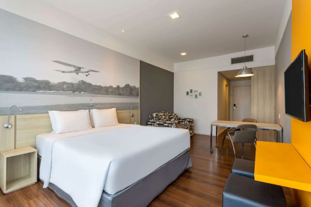 Quarto do Hotel Numaz Brás. Do lado direito dois puffs. uma televisãoe uma mesa de trabalho. Do lado esquerdo uma cama de casal, uma cômoda com abajur de cada lado e uma poltrona. Imagem para ilustrar post sobre hotéis perto do Expo Center Norte.