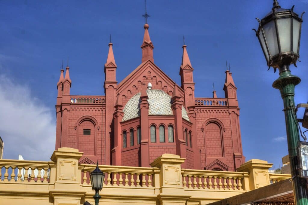 A imagem mostra a fachada do Centro Cultural Recoleta, um edifício icônico localizado no bairro da Recoleta, em Buenos Aires. A construção possui um estilo arquitetônico neogótico, caracterizado por suas torres pontiagudas e detalhes ornamentados. A cor predominante do edifício é um tom avermelhado, com uma cúpula metálica prateada no centro. À frente do edifício, há um parapeito em tom amarelo claro, complementando o contraste de cores com o azul do céu ao fundo. A lanterna decorativa no lado direito da imagem acrescenta um toque de elegância ao cenário urbano.