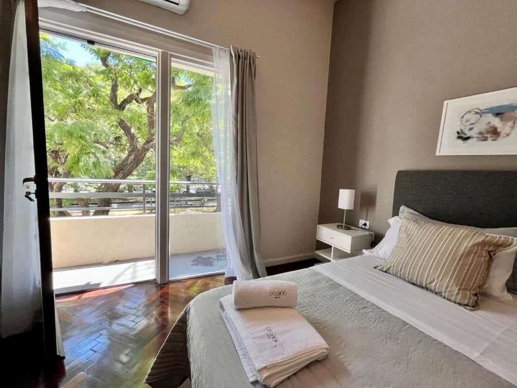 Uma cama bem arrumada ao lado de uma porta de vidro que dá para uma varanda com árvores visíveis do lado de fora, no DEL900 apartamentos, em Buenos Aires.