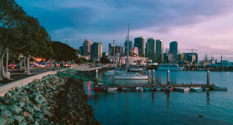 A foto mostra uma das marinas na baía de San Diego. Ela foi tirada ao anoitecer e mostra vários barcos atracados do lado esquerdo, perto das pedras. Do lado direito está a água e ao fundo os diversos prédios. A imagem ilustra o post de transportes em San Diego.