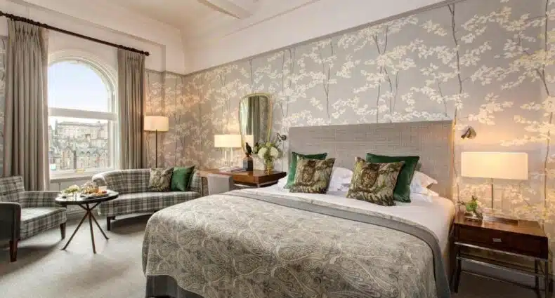 A foto mostra um quarto bem decorado com uma cama grande no centro, coberta com roupas de cama estampadas. Há dois criados-mudos ao lado da cama, cada um com um abajur. Em frente à cama, há duas poltronas estofadas com uma pequena mesa entre elas, exibindo o que parece ser uma bandeja de frutas e bebidas. O quarto tem paredes em tons neutros adornadas com papel de parede floral e uma grande janela que permite a entrada de luz natural. O ambiente geral é elegante e convidativo.