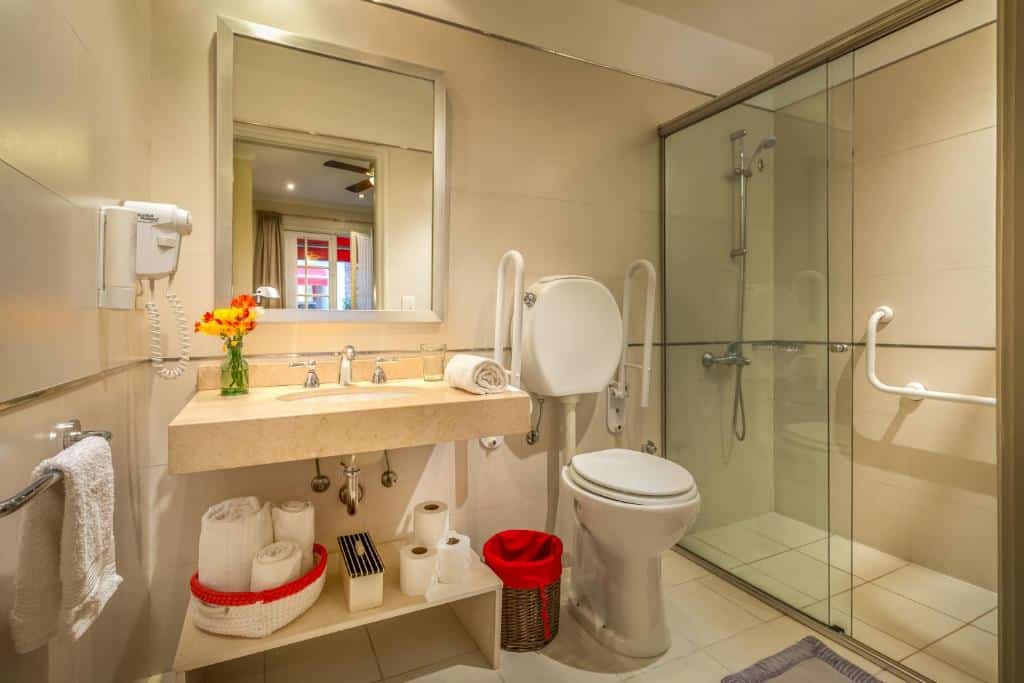 Banheiro acessível com barras de apoio, chuveiro com rodízio, vaso sanitário, pia, espelho, flores e toalhas em prateleiras abertas, no Duque Hotel Boutique & Spa.