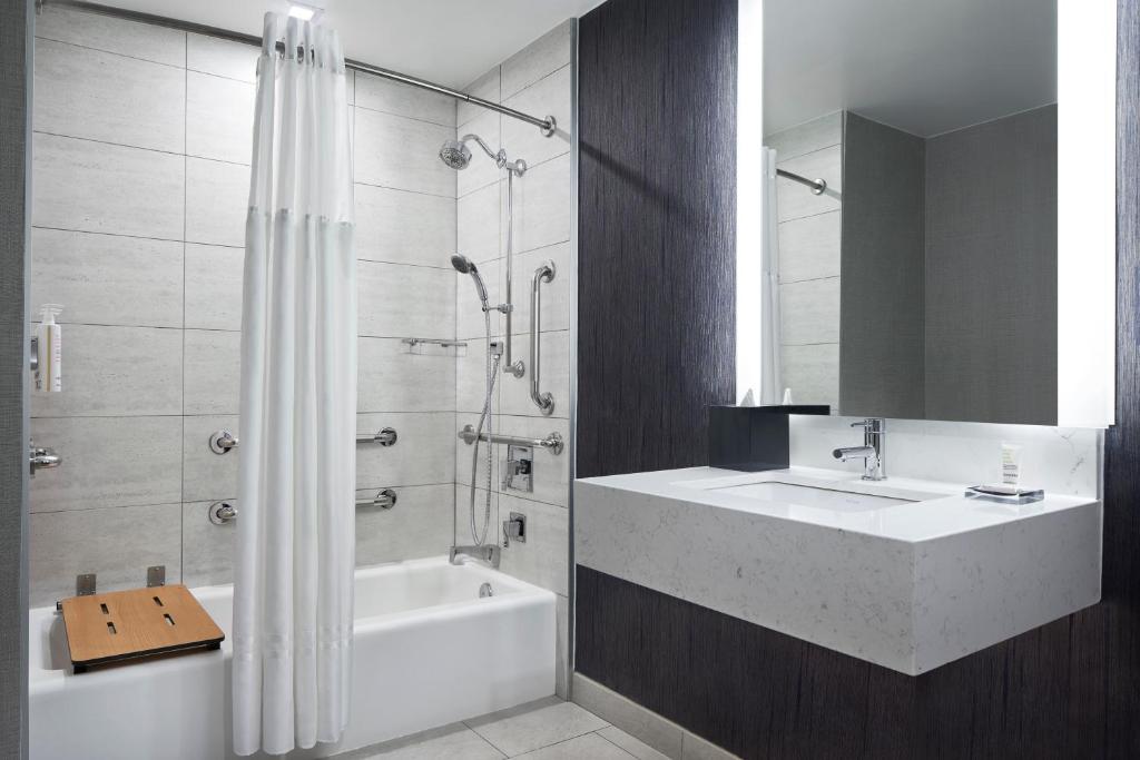 No banheiro do New York Marriott Marquis Hotel, há uma pia retangular branca com uma torneira moderna. À direita da pia, um espelho grande com iluminação embutida. À esquerda, há uma banheira com cortina de chuveiro branca. A parede do fundo é revestida com azulejos cinza.