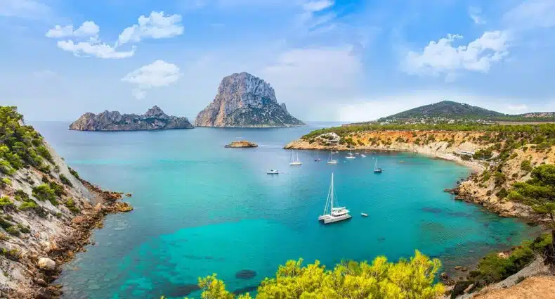 A imagem retrata uma deslumbrante baía em Ibiza, com águas azul-turquesa cristalinas, onde vários veleiros e iates estão ancorados tranquilamente para ilustrar o post sobre o que fazer em Ibiza. À esquerda e à direita, colinas cobertas por vegetação mediterrânea, principalmente pinheiros, descem em direção ao mar, enquadrando a baía. No centro da imagem, a icônica formação rochosa de Es Vedrà se ergue imponente do mar, criando um ponto focal dramático. O céu é azul claro com algumas nuvens brancas, completando o cenário paradisíaco e sereno.