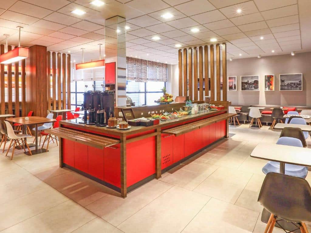 A imagem mostra a área do café da manhã do Ibis Buenos Aires Obelisco. O espaço é moderno e bem iluminado, com móveis contemporâneos. No centro, há uma ilha de buffet com um acabamento em vermelho, onde são dispostos itens como frutas, pães e bebidas. Ao redor, mesas e cadeiras coloridas oferecem assentos confortáveis para os hóspedes. A decoração é minimalista, com divisórias de madeira que separam o ambiente de maneira sutil, e fotos em preto e branco nas paredes, que complementam a estética clean do local.