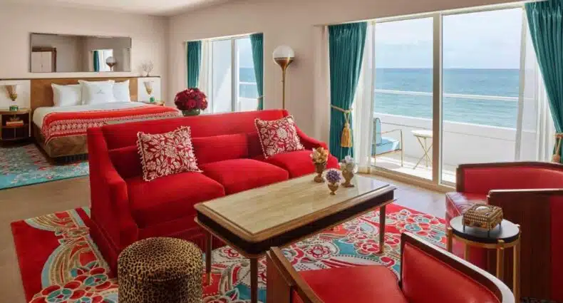 Suíte de hotel com esquema de cores vermelho e turquesa, com cama king size, vista para o mar, área de estar e janelas do chão ao teto.