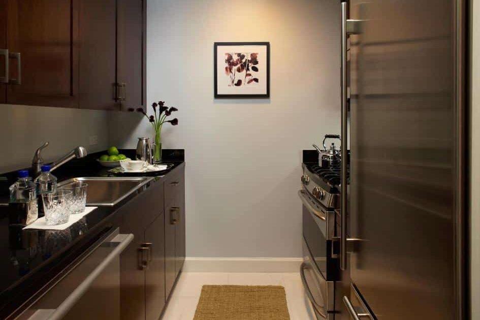 Na cozinha da suíte do Trump International Hotel há um balcão preto à esquerda com uma pia de aço inoxidável. Acima do balcão, armários de madeira escura. À direita, há um fogão e um forno embutidos. No canto direito, um refrigerador de aço inoxidável. Ao centro, há uma pequena mesa com uma fruteira e copos.