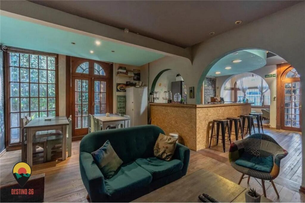 Lounge compartilhado do Destino26 Hostel, com sofás, mesa de centro, mesas e cadeiras e um bar. Imagem para ilustrar post sobre hostels em Montevidéu.