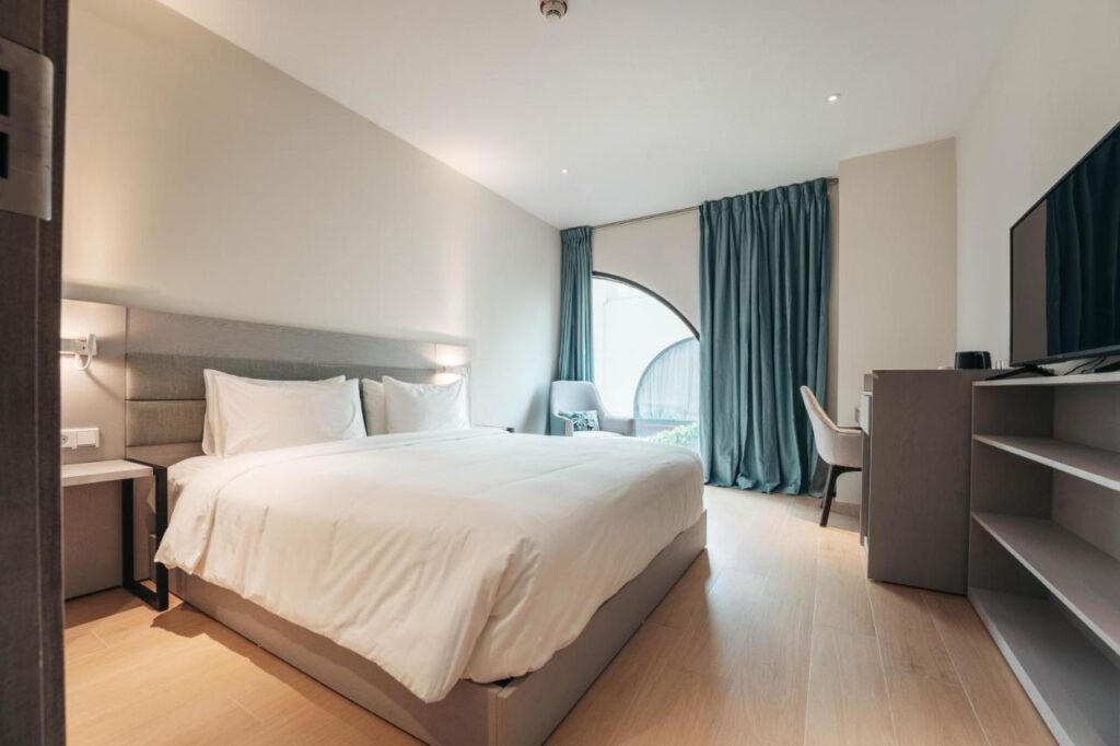 Quarto do Elements Hotel & Shops. Uma cama de casal no canto esquerdo. De frente uma cômoda, uma TV, uma mesa de trabalho. No fundo uma poltrona e uma janela.