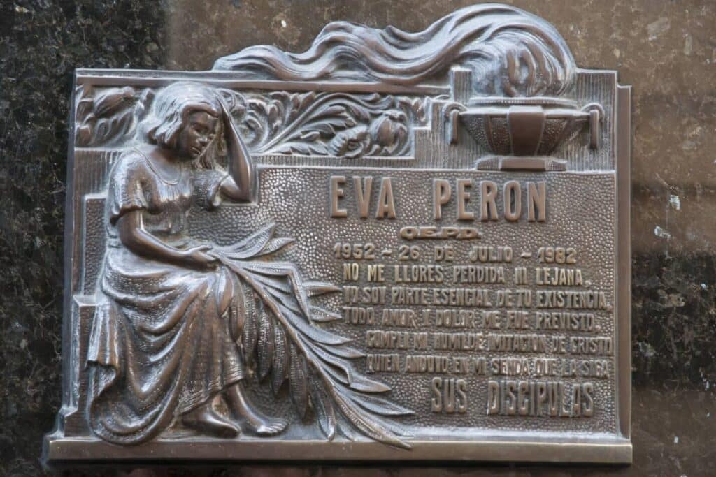 A imagem mostra uma placa de bronze com um epitáfio dedicado a Eva Perón, localizada no Cemitério da Recoleta em Buenos Aires. A placa apresenta uma representação em relevo de uma figura feminina sentada, com expressão triste, segurando folhas de palma, que simbolizam a vitória e a paz. Acima da figura, há uma chama eterna, representando a lembrança perpétua de Eva Perón. O texto na placa inclui seu nome, a data de sua morte (26 de julho de 1952), e uma mensagem que fala sobre a continuidade de sua essência e amor, mesmo após sua partida. É uma homenagem emocionada e reverente, colocada por suas discípulas.