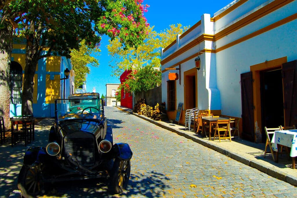 A imagem mostra uma rua charmosa e pitoresca em Colonia del Sacramento, uma cidade histórica localizada no Uruguai, conhecida por suas ruas de paralelepípedos e arquitetura colonial. No primeiro plano, há um carro antigo estacionado na rua, adicionando um toque nostálgico e vintage à cena. As fachadas das casas ao longo da rua são pintadas em cores vibrantes e contrastantes, com detalhes arquitetônicos que remetem ao período colonial. Árvores com folhagem densa e flores coloridas projetam sombras no chão, criando um jogo de luz e sombra que realça a beleza da paisagem urbana. Mesas e cadeiras de cafés ou restaurantes ao ar livre estão dispostas na calçada, sugerindo um ambiente acolhedor e convidativo para os visitantes. Ao fundo, a rua se estende em direção a uma torre distante, possivelmente uma igreja ou outro edifício histórico, completando a atmosfera tranquila e charmosa da cidade.