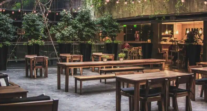 A imagem mostra a área externa do Anselmo Buenos Aires, Curio Collection by Hilton, um dos hotéis em San Telmo, Buenos Aires. O local conta com mesas e bancos de madeira dispostos em um ambiente cercado por plantas e luzes decorativas penduradas, criando uma atmosfera aconchegante e convidativa. Ao fundo, há uma parede de vidro que revela um espaço interno iluminado, provavelmente um café ou restaurante, que complementa a área externa, oferecendo um espaço perfeito para refeições ou relaxamento ao ar livre.