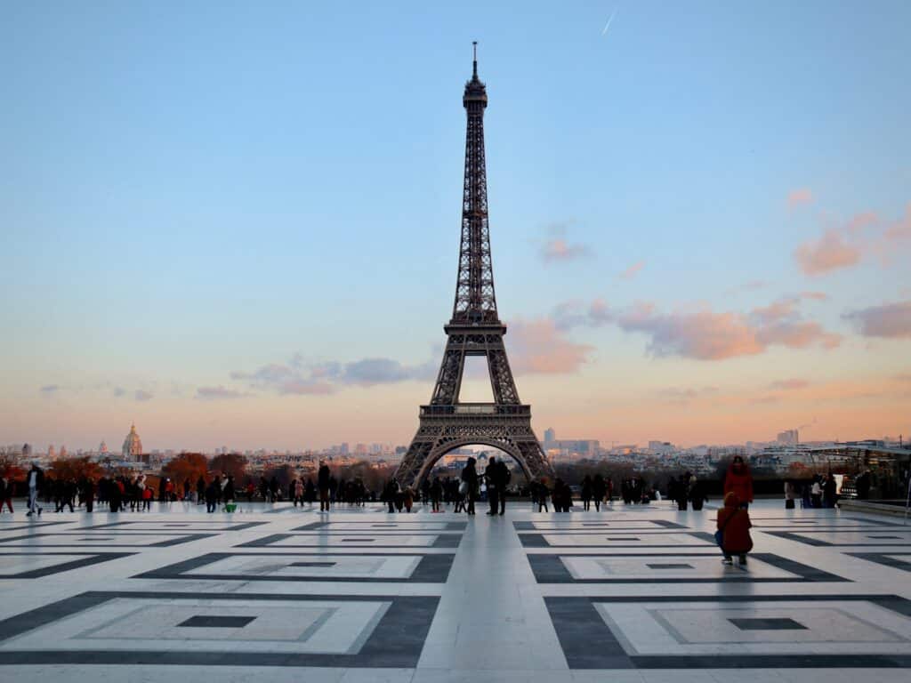 A imagem mostra a Torre Eiffel em Paris, vista de um amplo terraço com um padrão geométrico no chão. No horizonte, o céu está azul claro com algumas nuvens rosadas, sugerindo o final da tarde. Algumas pessoas estão caminhando ou tirando fotos, aproveitando a vista icônica. A torre, destacada no centro da imagem, contrasta com a suavidade do céu e a urbanização ao fundo.