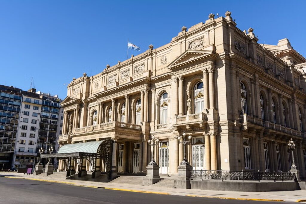A imagem mostra a imponente fachada do Teatro Colón, um dos edifícios mais emblemáticos de Buenos Aires. A arquitetura do teatro é grandiosa e clássica, com detalhes elaborados que refletem o estilo neoclássico. A fachada apresenta colunas robustas e arcos ornamentados, com grandes janelas e portas que reforçam a majestade do edifício. O Teatro Colón é reconhecido por sua importância histórica e cultural, sendo um dos principais locais de apresentações de ópera, balé e concertos na cidade.