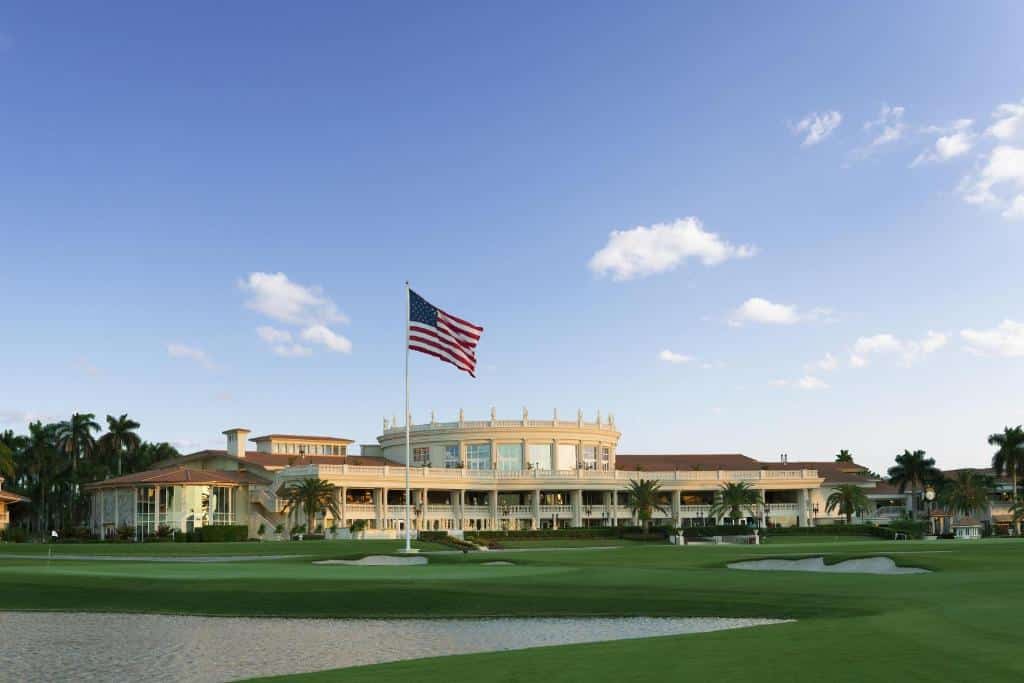 Imagem da frente do Trump National Doral Golf Resort com uma bandeira americana tremulando na frente, cercado por palmeiras e um campo de golfe bem conservado sob um céu azul.