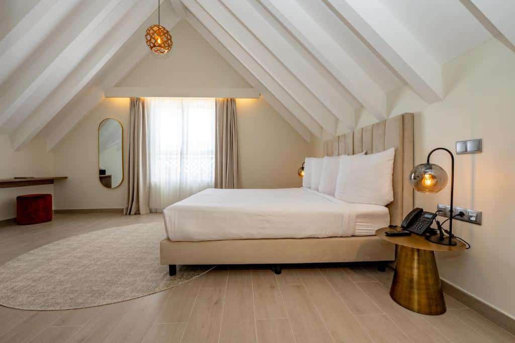 Quarto do Harbor Hotel & Casino. Uma cama de casal do lado direito, com uma mesa de apoio e abajur de cada lado. Do lado esquerdo uma mesa e um espelho. No fundo a janela.