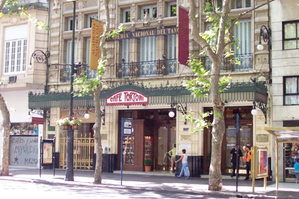 A imagem mostra a fachada do famoso Café Tortoni, localizado em Buenos Aires. O edifício tem uma arquitetura clássica, com detalhes ornamentais e janelas grandes. A entrada do café é destacada por um toldo verde com o nome "Café Tortoni" em letras vermelhas. Acima da entrada, há um letreiro indicando "Academia Nacional del Tango", o que realça a importância cultural do local. Árvores estão posicionadas em frente ao café, e é possível ver algumas pessoas caminhando na calçada, contribuindo para o clima movimentado e histórico da área.