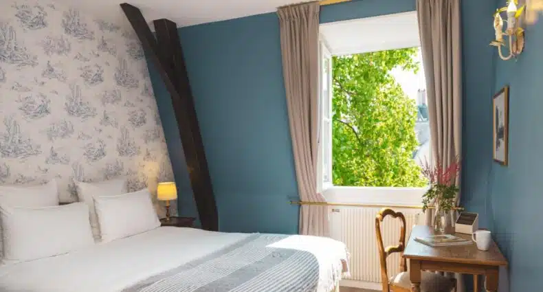 A imagem mostra um quarto do Hotel des Grandes Ecoles com uma cama de casal coberta com roupa de cama branca e um cobertor cinza listrado. A parede ao fundo tem um papel de parede decorado com padrões azuis e brancos, e uma janela aberta à direita permite a entrada de luz natural, revelando uma vista verdejante de árvores. Há uma mesa de madeira ao lado da janela, com uma cadeira, uma xícara e um vaso de flores. O ambiente é aconchegante e iluminado, e o hotel faz parte da lista de hotéis 3 estrelas em Paris.
