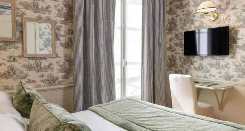 Quarto do Hotel du Champ de Mars, um dos hotéis com custo-benefício em Paris. A cama é coberta com colchas e almofadas em tons de verde suave. As paredes são revestidas com um papel de parede em estilo toile de jouy, apresentando cenas campestres em tons de cinza sobre um fundo bege. Uma TV de tela plana está montada na parede, acima de uma pequena escrivaninha com uma cadeira clara. As cortinas listradas em cinza e branco enquadram uma janela, permitindo a entrada de luz natural e criando um ambiente acolhedor e sereno.