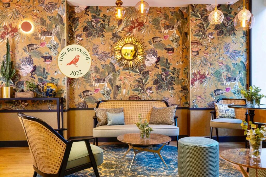 A imagem mostra uma área de lounge decorada de forma vibrante e acolhedora no Hôtel Jardin de Cluny. As paredes têm papel de parede com um design colorido de animais e plantas tropicais. Há um espelho dourado em forma de sol na parede central. O mobiliário inclui um sofá e cadeiras com estofamento claro e detalhes de vime, além de várias almofadas. No centro, há uma mesa de café redonda com um vaso de flores. A iluminação é suave, com luminárias pendentes de vidro e uma luz de parede circular. A decoração cria um ambiente elegante e confortável.
