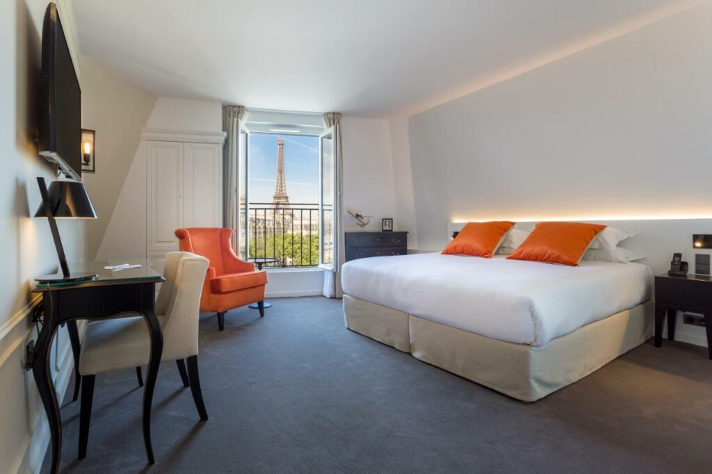 A imagem mostra um quarto elegante e moderno no Hôtel La Comtesse, um dos hotéis 4 estrelas em Paris, com uma cama grande coberta por lençóis brancos e almofadas laranja. Ao lado da cama, há mesas de cabeceira escuras e um telefone. Uma cadeira laranja está posicionada próxima à janela, que oferece uma vista deslumbrante da Torre Eiffel ao fundo. A parede ao lado da janela é branca, contrastando com o tapete cinza no chão. Na frente da cama, há uma escrivaninha preta com uma cadeira bege, e uma TV montada na parede acima dela.