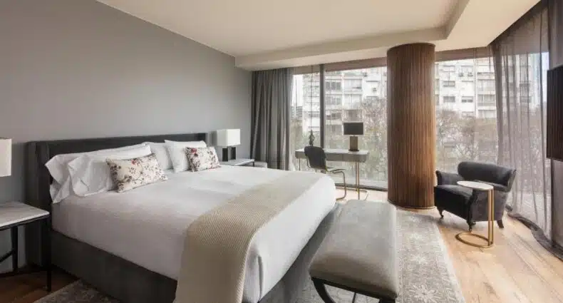 Quarto do Leading Hotels of the World. Uma cama de casal do lado esquerdo, na frente um sofá. No fundouma mesa de trabalho e uma poltrona e paredes de vidro. Imagem para ilustrar post sobre onde ficar em Montevidéu.