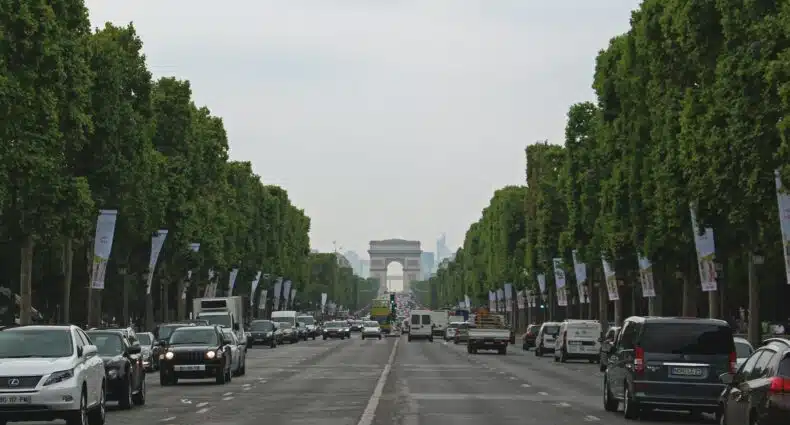 A imagem mostra uma ampla avenida ladeada por árvores verdes, com carros circulando em ambos os sentidos. Ao fundo, destaca-se o Arco do Triunfo, um famoso monumento parisiense. A avenida é a Champs-Élysées, uma das mais conhecidas de Paris, e está coberta por um céu nublado, criando uma atmosfera tranquila e típica de dias em que o sol está encoberto. Banners publicitários estão pendurados ao longo da avenida, adicionando cor ao cenário.