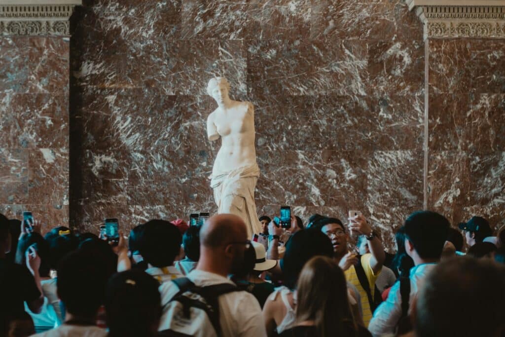 A imagem mostra uma estátua famosa, a Vênus de Milo, cercada por uma grande multidão de pessoas no Museu do Louvre. A estátua é de uma figura feminina sem braços, feita de mármore branco, destacando-se contra uma parede de mármore escuro. As pessoas ao redor estão tirando fotos com seus celulares, admirando a obra de arte. O ambiente está cheio, com muitas cabeças visíveis na parte inferior da imagem.