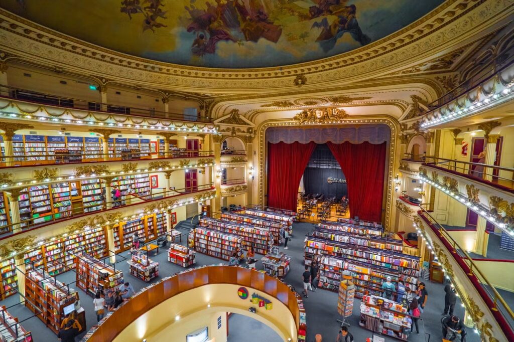 A imagem mostra o interior do El Ateneo Grand Splendid, uma das livrarias mais famosas e belas de Buenos Aires, Argentina. Originalmente construído como um teatro em 1919, o edifício mantém grande parte de sua arquitetura original, incluindo os balcões, o teto decorado com afrescos e o palco, que agora abriga um café. As estantes de livros ocupam o lugar onde antes ficavam as cadeiras do teatro, criando uma atmosfera única que mistura a grandiosidade de um espaço teatral com a tranquilidade de uma livraria. As luzes suaves e a decoração detalhada realçam a elegância do local, fazendo desta livraria um ponto turístico imperdível na cidade.