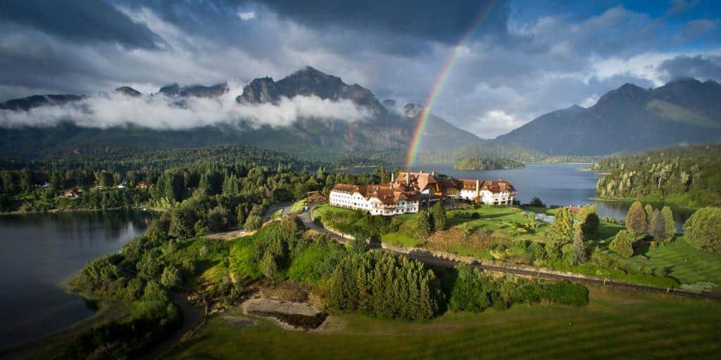 vista aerea do hotel Llao Llao em Bariloche, mostrando as montanhas ao fundo, com várias nuvens cobrindo seus picos, um belo lago aos pés das montanhas, com muitas árvores e pinheiros, e uma construção de paredes brancas, com diversas janelas e prédios bem no centro da imagem, com uma estrada de asfalto percorrendo a lateral do hotel e adentrando na mata.