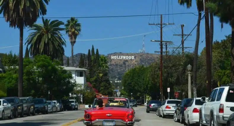A foto mostra um carro vermelho andando pelas ruas de Beverly Hills. Ao redor, estão diversas árvores e carros. Ao fundo, de frente para o veículo, está a placa de Hollywood vista de longe. A foto é capa do post de transportes em Los Angeles.