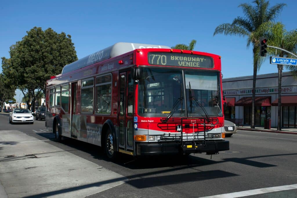 Esse é um dos ônibus de Los Angeles. Ele é todo vermelho e está escrito "Broadway - Venice". Ao redor estão estabelecimentos, árvores e alguns carros.