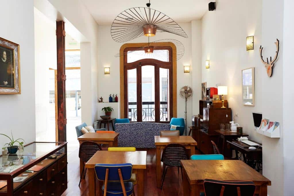 A imagem mostra uma área comum do L'Adresse Hôtel Boutique, decorada com um estilo que combina o clássico e o moderno. O ambiente é iluminado naturalmente através de uma grande porta de madeira com vidro, que dá acesso a uma sacada. As paredes brancas são complementadas por detalhes decorativos, como quadros e luminárias de parede. Há uma variedade de móveis, incluindo mesas de madeira com cadeiras coloridas, um sofá com estofado azul e cadeiras de design contemporâneo. Um grande lustre de arame pendente adiciona um toque moderno ao ambiente, enquanto móveis antigos, como um armário e uma escrivaninha, contribuem para um charme vintage. O espaço parece acolhedor e bem organizado, ideal para momentos de relaxamento ou socialização.