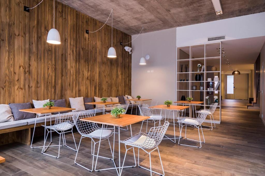 A imagem mostra uma área de refeições elegante e moderna do Lemon Suites. O espaço é decorado com paredes revestidas de madeira, que adicionam um toque acolhedor ao ambiente. As mesas são de madeira clara, combinando com o revestimento das paredes, e são acompanhadas por cadeiras de metal com design moderno e minimalista. Almofadas em tons neutros estão dispostas ao longo do banco fixo na parede, proporcionando conforto adicional. As luminárias pendentes brancas oferecem iluminação suave, criando uma atmosfera agradável. Ao fundo, há uma estante decorativa que separa a área de refeições de outro ambiente, contribuindo para a organização e o estilo do espaço.