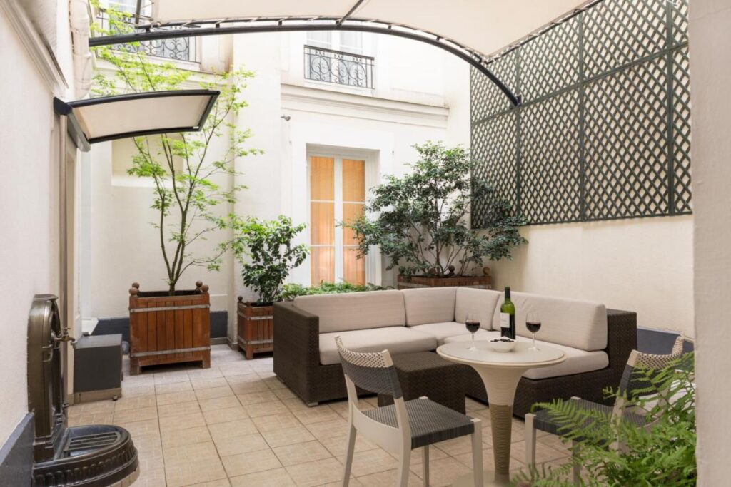 A imagem mostra um pátio externo pequeno e aconchegante no Magda Champs Elysées, decorado com plantas em vasos de madeira e uma treliça na parede, que dá um toque de jardim urbano. Há um sofá de canto em tons de bege e marrom escuro, criando um espaço confortável para sentar. Em frente ao sofá, há uma mesa redonda com duas cadeiras, sobre a qual estão uma garrafa de vinho e duas taças. O ambiente é coberto por uma estrutura leve que proporciona sombra, tornando o espaço ideal para relaxar ao ar livre em um ambiente privado e tranquilo.