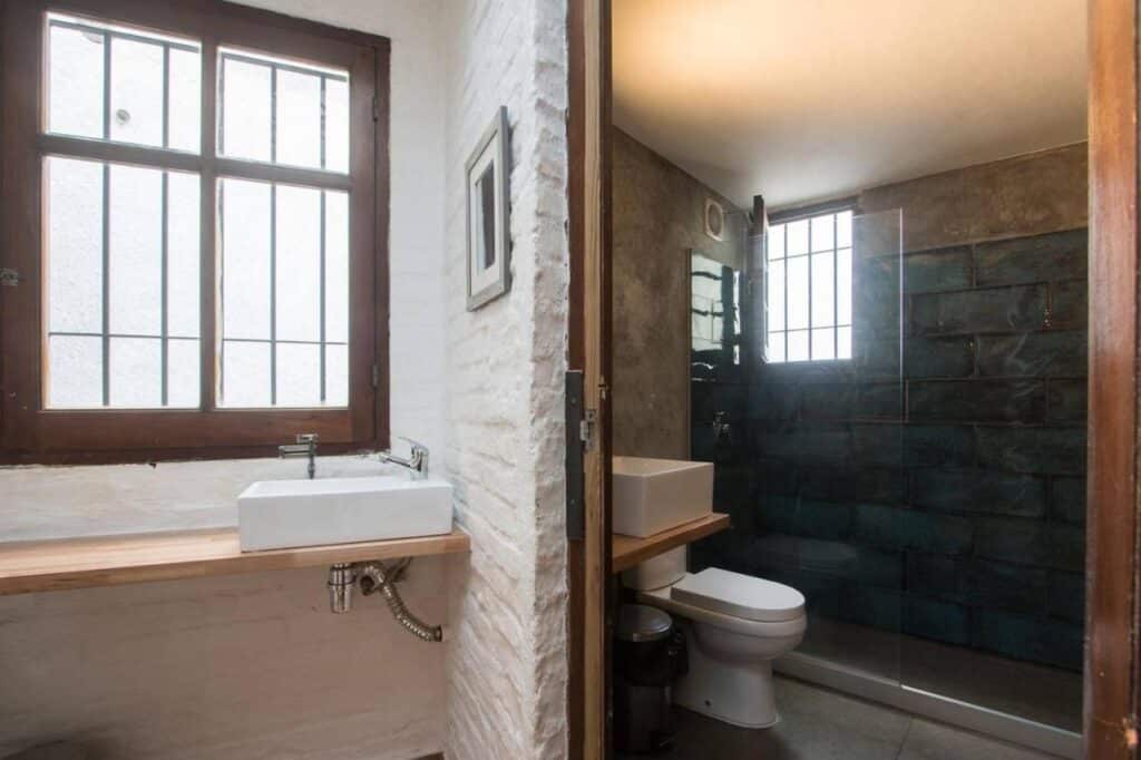 Banheiro do MedioMundo Hostel. Uma pia e uma janela do lado esquerdo, do lado dirieto outra pia, um vaso sanitário e o box.